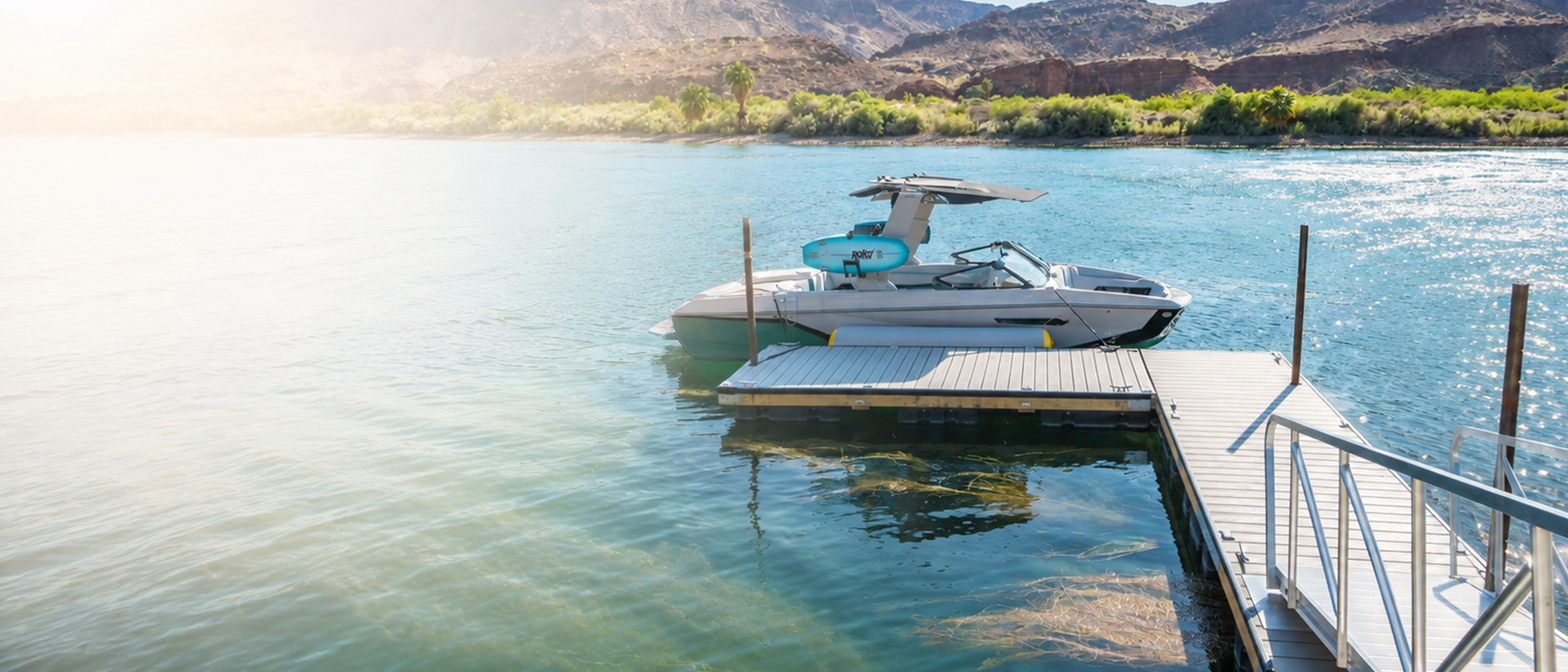 A wakeboat moored at a floating dock on a turquoise desert lake, red rock canyon rising behind it